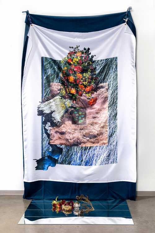 A tapestry showing a dead tree stump, bright, lively flowers and pebbles. Below on a mirror sits still-life elements reflecting the elements in the tapestry