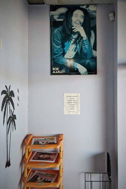 A blue monochrome poster of Bob Marley hangs on a lilac wall. Beneath the poster is a sign that reads "In practice with social distancing please keep 2 meters (6 feet) apart. Thank you." Below sits a yellow worn out plastic shelf housing magazines, and a balvk chair frame next to it. The wall on the left has a black decal of palm trees.